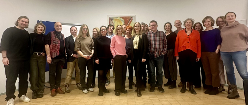 Gruppe von rund 20 Personen bei einem Treffen von Fachgesellschaften in einem Innenraum, stehend in einer Reihe, einige in der Mitte hervorgehoben, freundliche Atmosphäre, Holzfußboden und Kunstbild im Hintergrund.