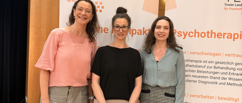 Drei Frauen stehen vor Roll-ups des Tiroler Landesverbands für Psychotherapie bei einem Psyche Kompakt Vortrag, freundlich in die Kamera blickend, Innenraum mit Holzverkleidung.
