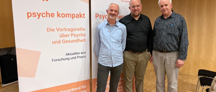 Drei Männer stehen vor Roll-up-Bannern des Tiroler Landesverbands für Psychotherapie mit der Aufschrift "Psyche kompakt – Die Vortragsreihe über Psyche und Gesundheit" in einem Veranstaltungsraum.